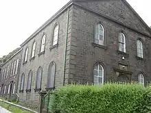 A two-storey gabled stone building seen from an angle with another building attached behind it. There are round-headed windows and a door in the nearer building with straight-headed windows in the building behind it.