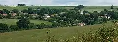 The roofs of many houses, and a church spire can be seen in a green valley with several trees.