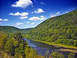 A river curves between two forested mountains under a bright blue sky with a few clouds