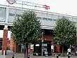 A brown-bricked building with a large, grey sign reading "WEST HAM" in white letters and four people in front all under a light grey sky