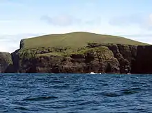 A green hill ends in dark brown cliffs over blue water.