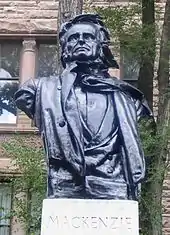 A black bust of Mackenzie looking upwards. The statue is placed on a white stone stand engraved with the word "Mackenzie"