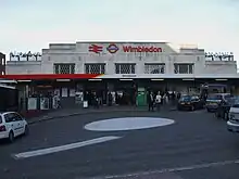 A white-bricked building with a sign reading "Wimbledon" in red letters and people walking in front all under a light blue sky with white clouds