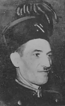 Head and shoulders shot of a thin faced man with a toothbrush moustache, wearing military uniform style dress: pillbox hat with plumes and insignia and buttoned up jacket with lapel pickaxe insignia.