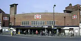 A brown-bricked building with a rectangular, dark blue sign reading "WOOD GREEN STATION" in white letters all under a light blue sky