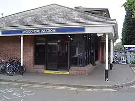 A red-bricked building with a rectangular, dark blue sign reading "WOODFORD STATION" in white letters all under a clear, white sky