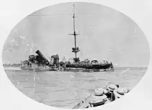 A badly damaged ship sits in the water, while in the foreground sailors in another vessel watch on.