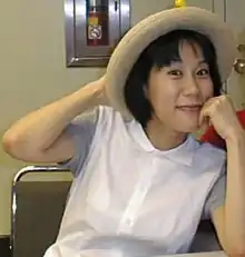 A seated Yoko Kanno, wearing a hat and smiling at the camera