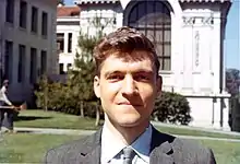 A man in a suit faces the camera while he stands in front of a building.